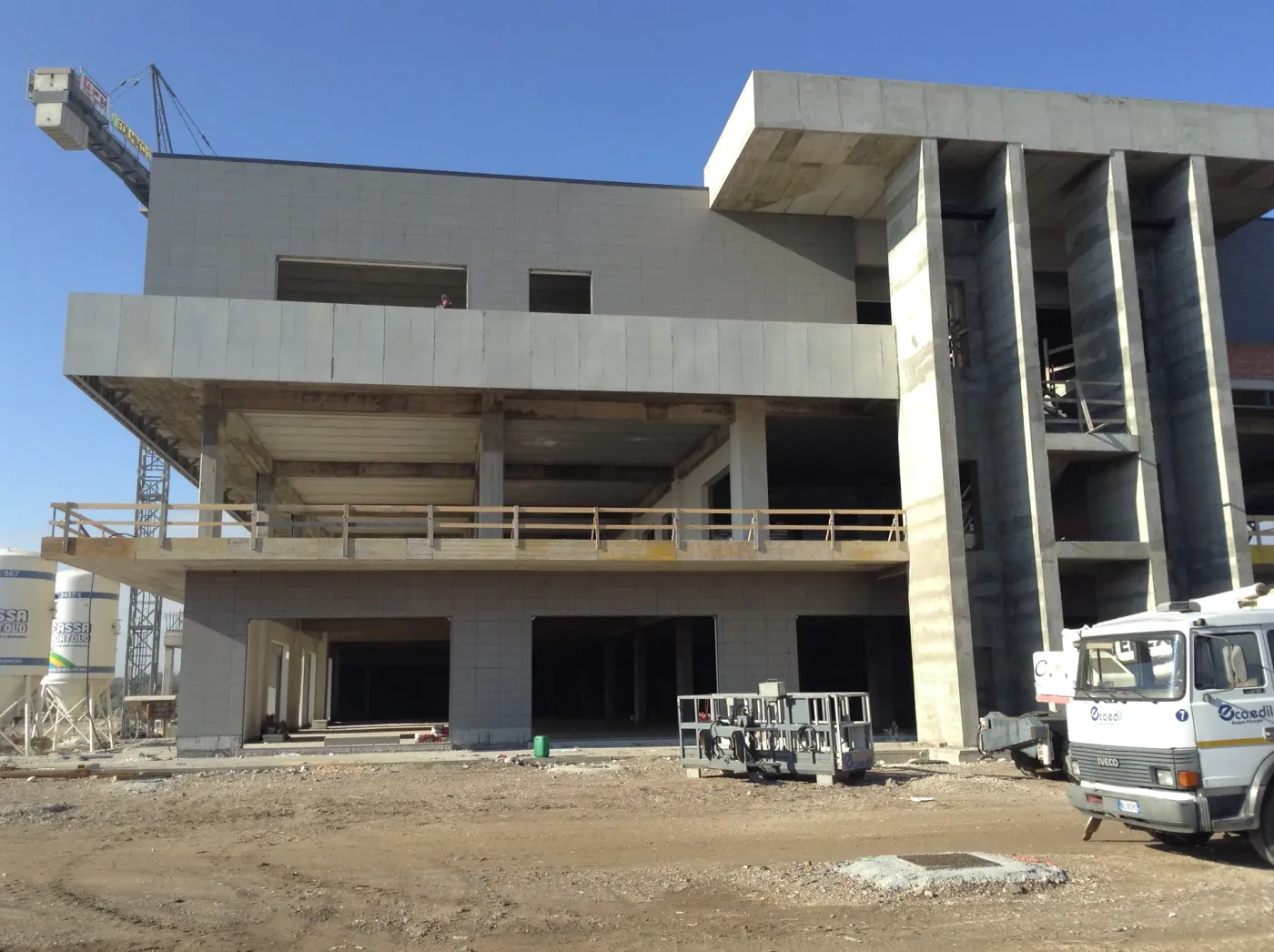 Fronte dell’edificio con pilastri in cemento armato a tutta altezza e autocarro di cantiere.